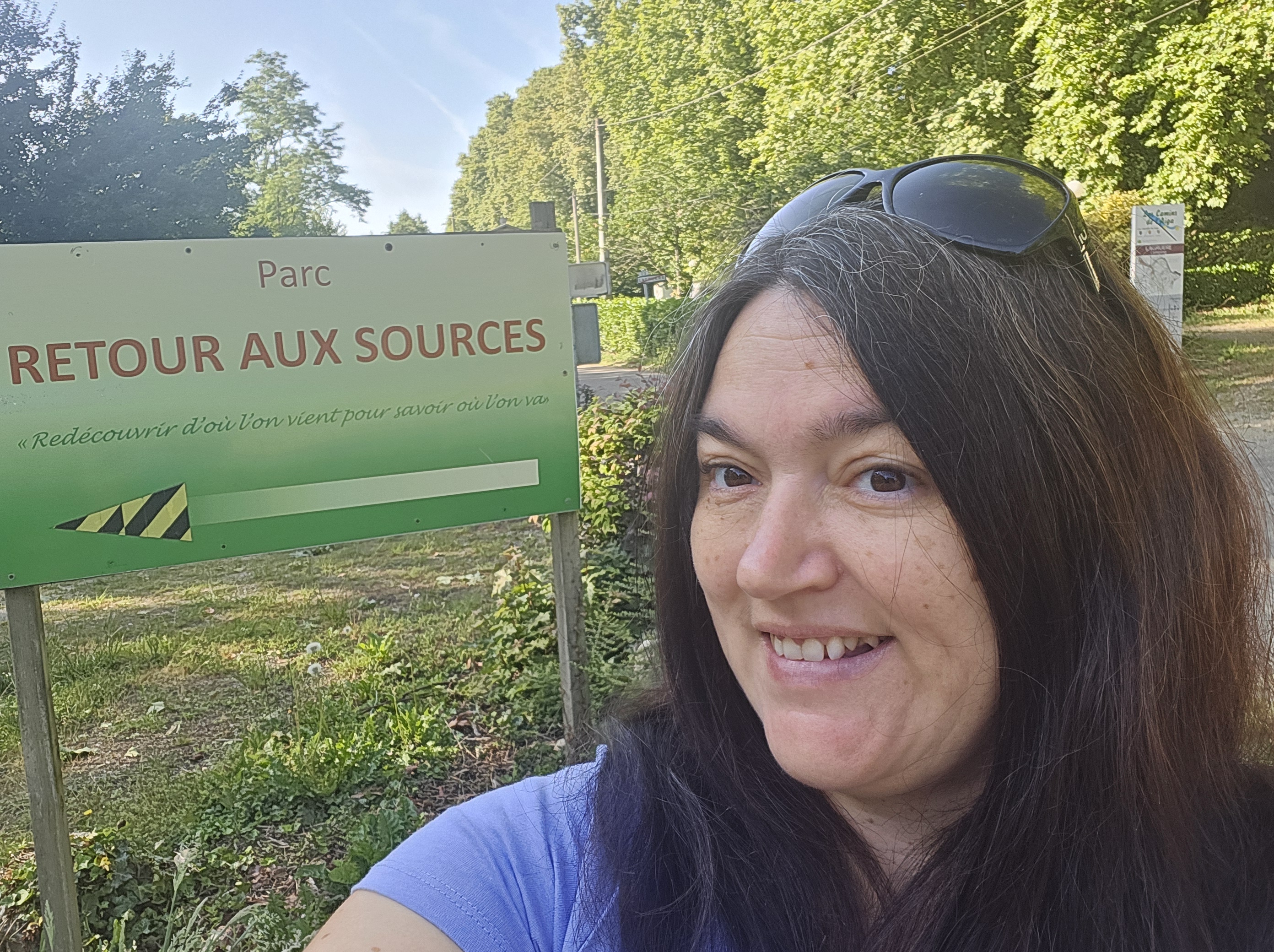  Gaëlle, conseillère bien-être, debout à l’entrée du Parc Retour aux Sources à Lagruère. Elle accueille les visiteurs avec un sourire chaleureux, entourée de verdure, symbolisant la reconnexion à la nature et à soi-même. L’environnement paisible du parc reflète l’esprit de l’expérience "L’Essentiel en Forêt" : calme, ancrage, et ressourcement. Une lumière douce filtre à travers les arbres, évoquant un moment suspendu hors du temps.